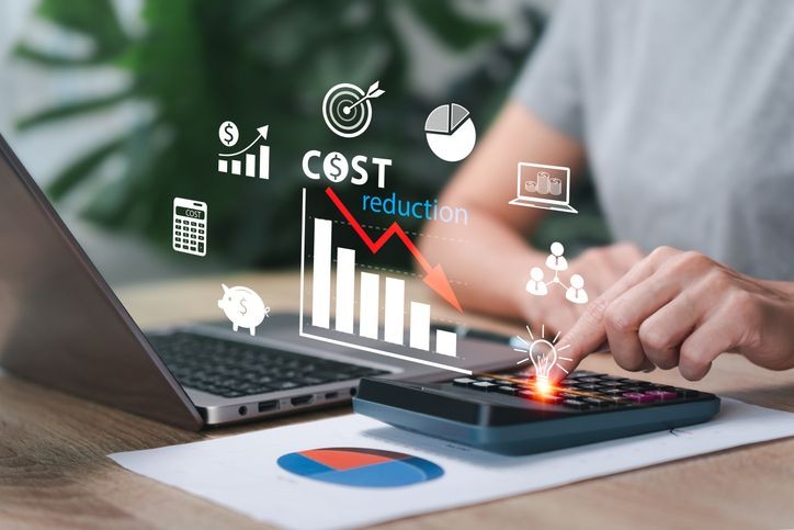 Person using a calculator with cost reduction graphics and charts near a laptop on a desk.