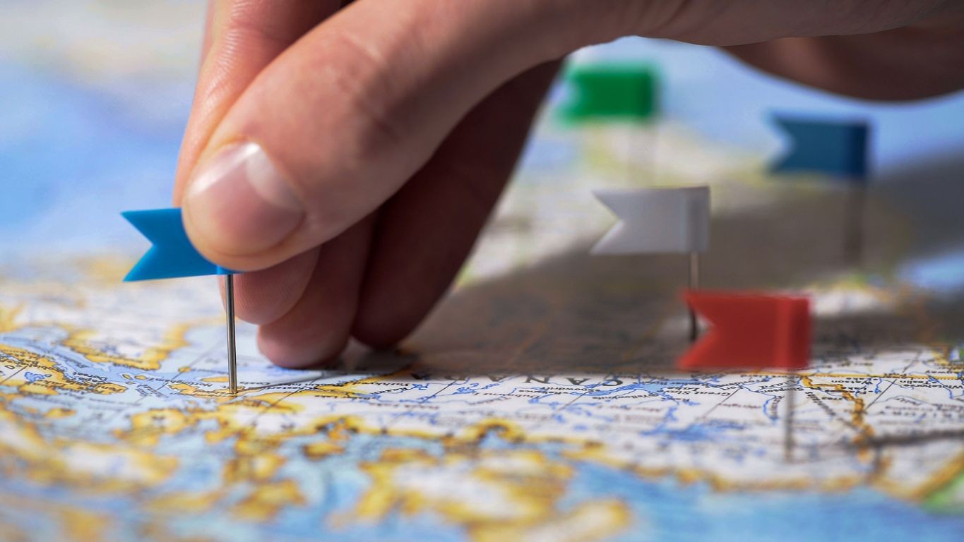 Hand placing a blue flag pin on a map with other colored pins visible in the background.