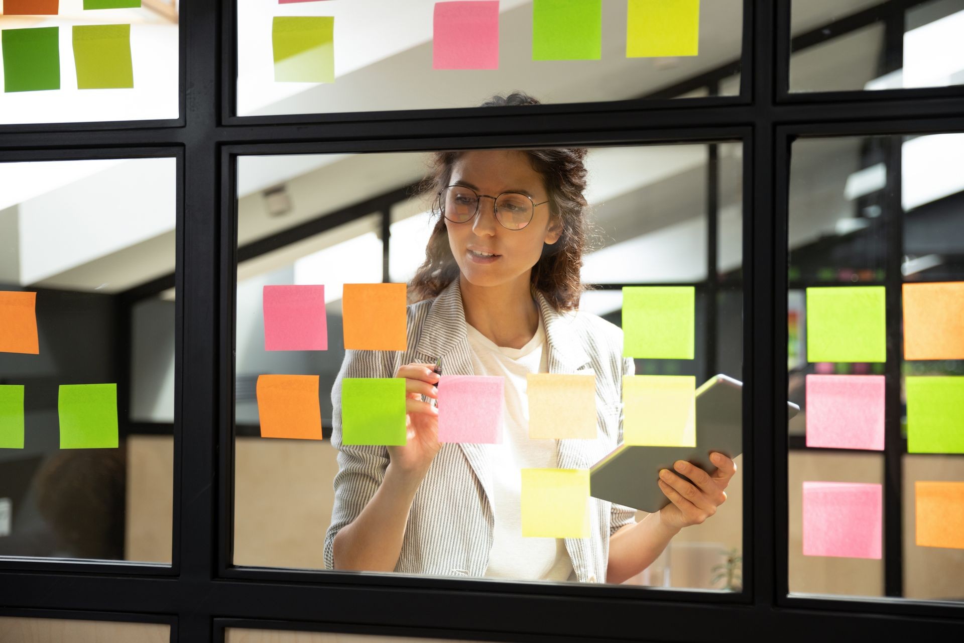 Person organizing colorful sticky notes on a glass wall while holding a tablet in an office setting.